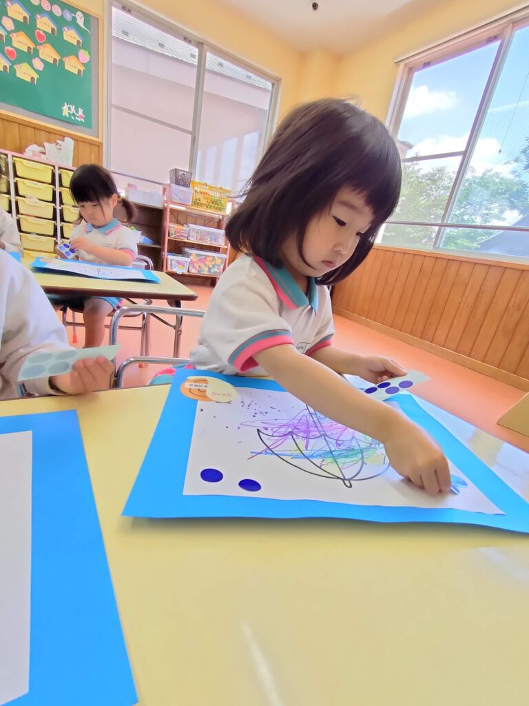 「ここに決めた」——青いシールを雨粒に見立てて貼るいちご組(2歳児クラス)